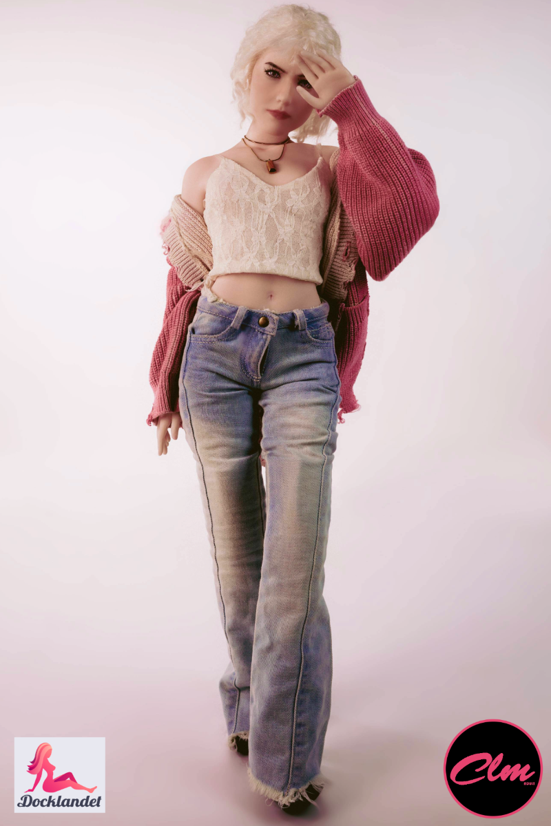 poupée sexuelle Mini poupée blonde réaliste en silicone de 60 cm Tara en jean, haut court et veste rose, debout sur fond clair de studio.